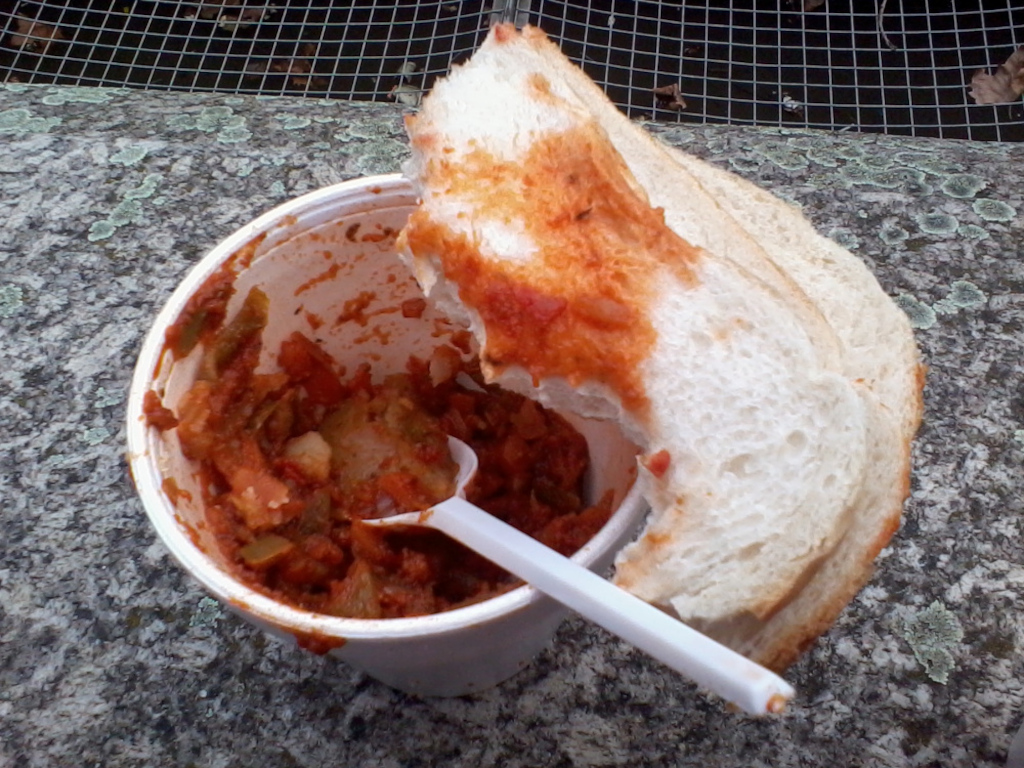 Partially eaten meal consisting of a stew-like dish in a white disposable cup, and a piece of white bread that has been used to scoop the stew. The bread is significantly soaked with the red sauce of the stew. The meal is sitting on a mottled grey stone surface. A plastic spoon is also visible, partially inserted into the cup. The background includes a metal mesh fence.