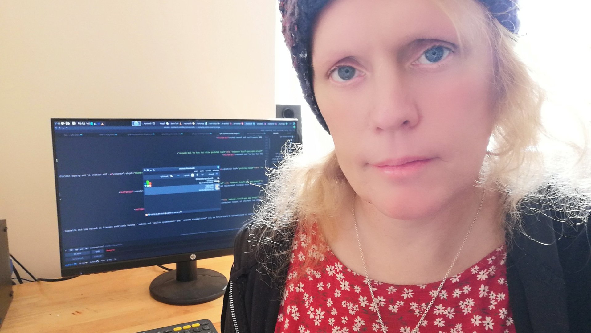 Selfie of Léonie, taken from the chest up, with a computer monitor showing code in the background. The woman is wearing a red floral top, a dark jacket, and a knitted hat. Her expression is serious and somewhat pensive. The computer screen shows what appears to be lines of code, suggesting a technological or programming context. The overall impression is of a woman focused on her work.