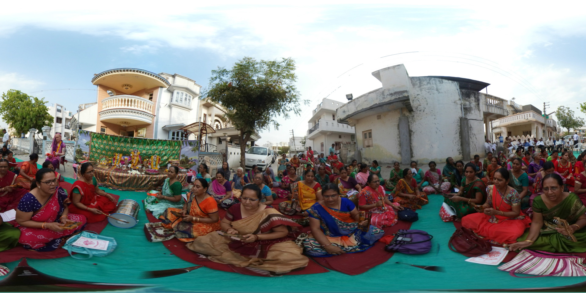 Large gathering of predominantly women, mostly middle-aged and older, seated on a vibrant teal-green tarp spread across a street. They are dressed in a kaleidoscope of vibrant South Asian saris and clothing, with rich jewel tones of red, orange, pink, purple, green, and gold predominating. Many women hold small objects, possibly prayer items or musical instruments. Some are actively engaged in what appears to be a religious ceremony, while others seem to be observing. In the foreground, several women are prominently featured; one, in a deep red sari, holds what looks like a small percussion instrument. Another, in a beige and gold sari, is seated centrally, appearing calmly observant. A few women in the background are dressed in lighter-colored clothing, standing or sitting slightly apart. A small shrine or altar with religious images and offerings is visible behind the seated women. A dholak (a double-sided barrel drum) rests on the ground near the central figures. A few men are visible in the distance at the edges of the scene, observing. The photograph employs a 360-degree panoramic perspective, encompassing a wide field of view. This offers a comprehensive view of the entire scene, placing the viewer at the center of the gathering. The circular frame accentuates the communal nature of the event. The subjects are arranged in a semi-circular formation around the central altar, creating a natural focal point.