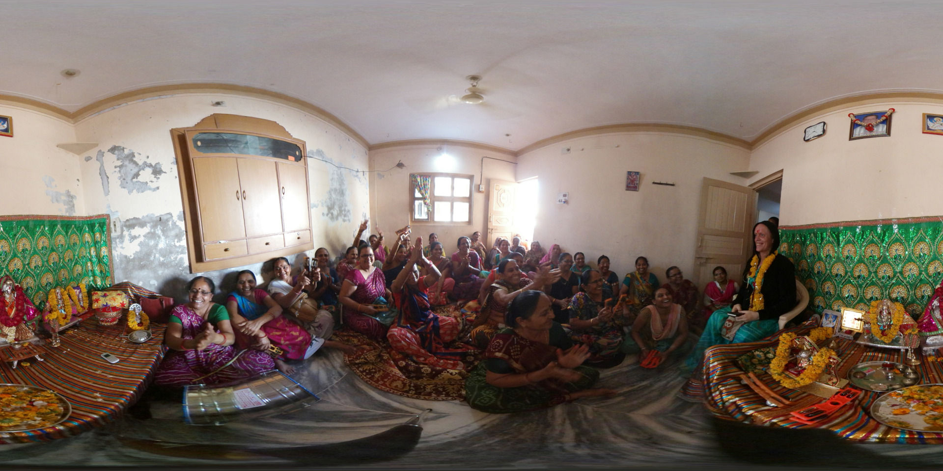360 degree panoramic view, dominated by a large group of women, predominantly of South Asian descent, gathered in a room. They are mostly seated on colorful rugs spread across the floor, many clapping, smiling, and gesturing excitedly. Their clothing is a vibrant mix of brightly colored saris, blouses, and other traditional garments, with a rich tapestry of textures and patterns. Leonie, appearing to be a Westerner, sits slightly apart, wearing a teal and black outfit and a garland, calmly observing the group. She is holding what seems to be a smartphone. Two smaller shrines are visible, one at each end of the rug, adorned with colourful flowers, offerings, and small statues.