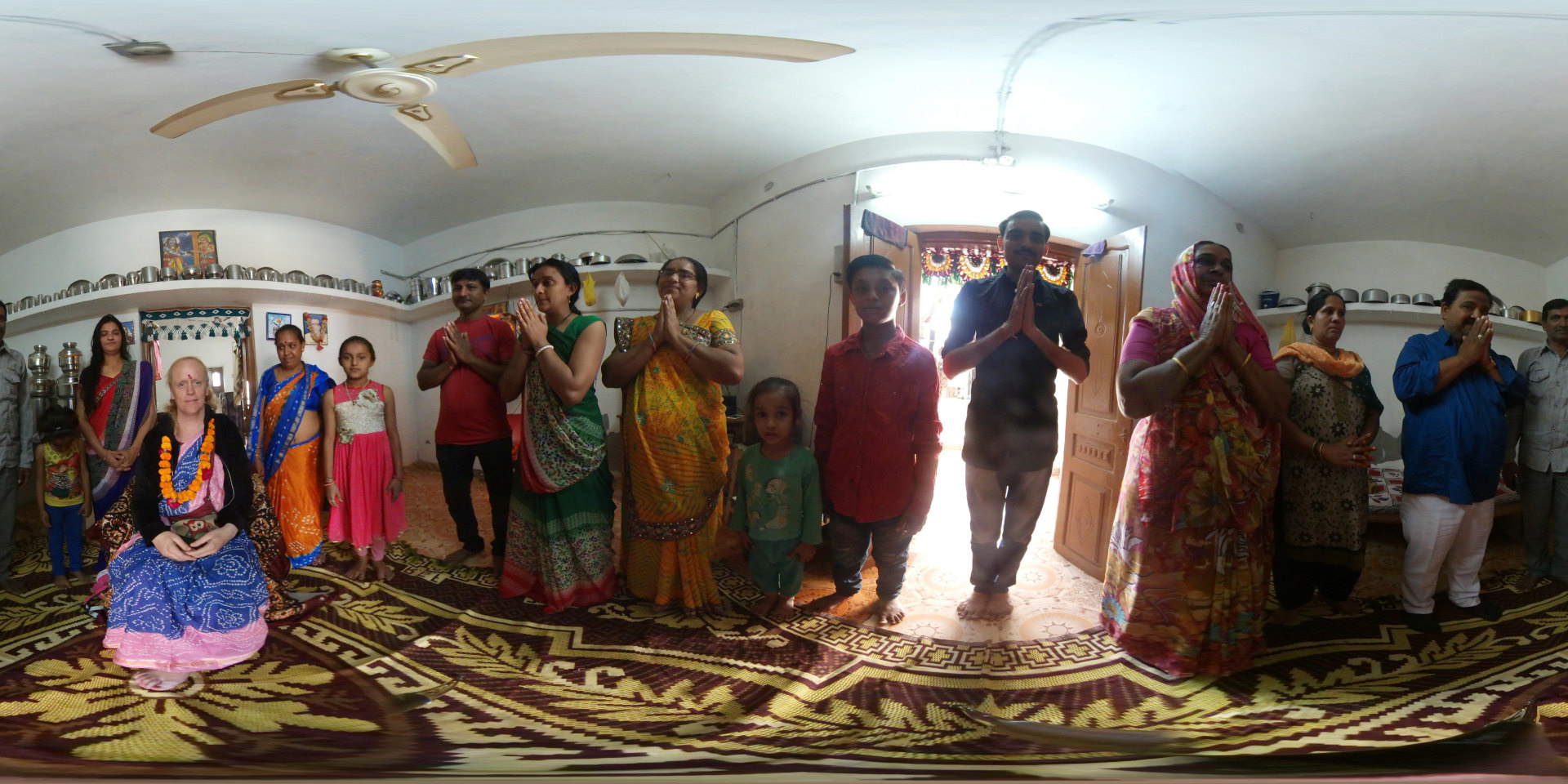 360 degree panoramic view, dominated by a large group of women, predominantly of South Asian descent, gathered in a room. They are mostly seated on colorful rugs spread across the floor, many clapping, smiling, and gesturing excitedly. Their clothing is a vibrant mix of brightly colored saris, blouses, and other traditional garments, with a rich tapestry of textures and patterns. Leonie, appearing to be a Westerner, sits slightly apart, wearing a teal and black outfit and a garland, calmly observing the group. She is holding what seems to be a smartphone. Two smaller shrines are visible, one at each end of the rug, adorned with colourful flowers, offerings, and small statues.