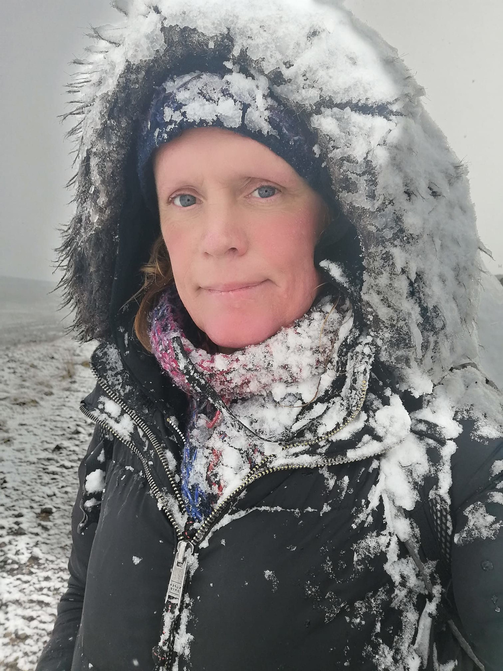 Close-up of a Leonie's face and upper body, heavily covered in snow. She's wearing a dark-coloured hooded jacket with fur trim, a scarf, and a hat, all visibly coated in snow. Her expression is neutral, almost stoic. The background is blurred but shows a snowy, outdoor landscape. The overall impression is one of cold weather and exposure to the elements.