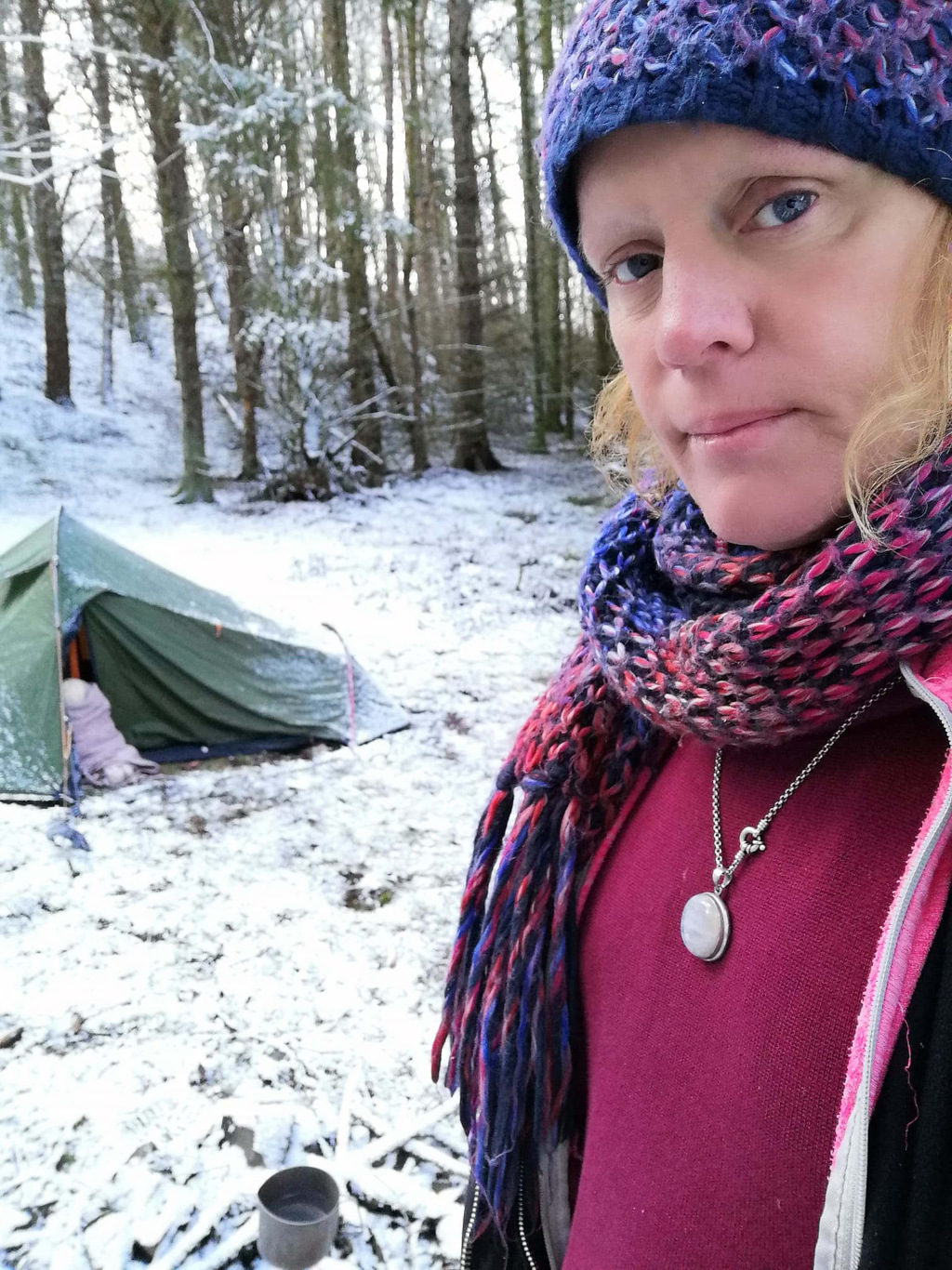 Leonie standing in a snowy woodland setting, in front of a small green tent. She is wearing a knitted hat and scarf, a maroon top, and a necklace with a pendant. Her expression appears serious or perhaps pensive. The overall atmosphere is one of quiet solitude in a wintery, natural environment.