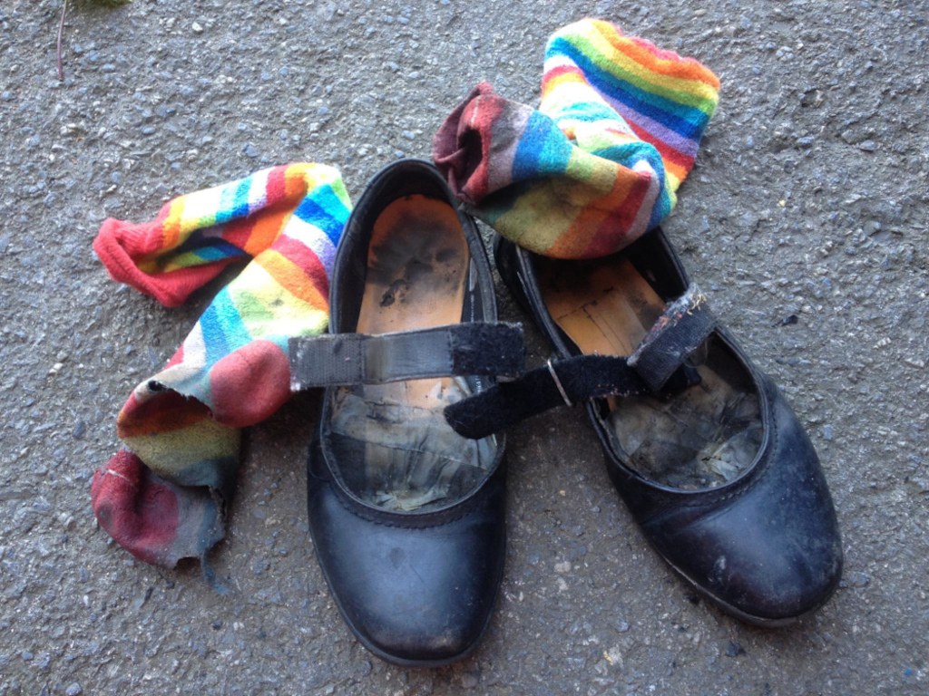 Worn-out black Mary Jane shoes and a colourful, striped sock lying on a gray asphalt surface. The sock is partially torn and dirty, reflecting wear and tear. The shoes are also visibly used and dirty, showing signs of significant wear. The overall impression is one of abandonment or neglect.
