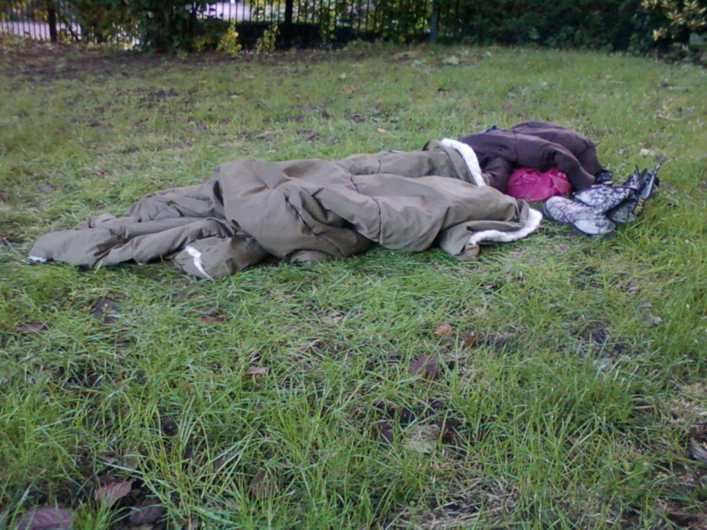 Sleeping outdoors, wrapped in a sleeping bag on a grassy area. Near the sleeping bag are some clothes and footwear. The overall impression is of someone sleeping rough, possibly homeless.