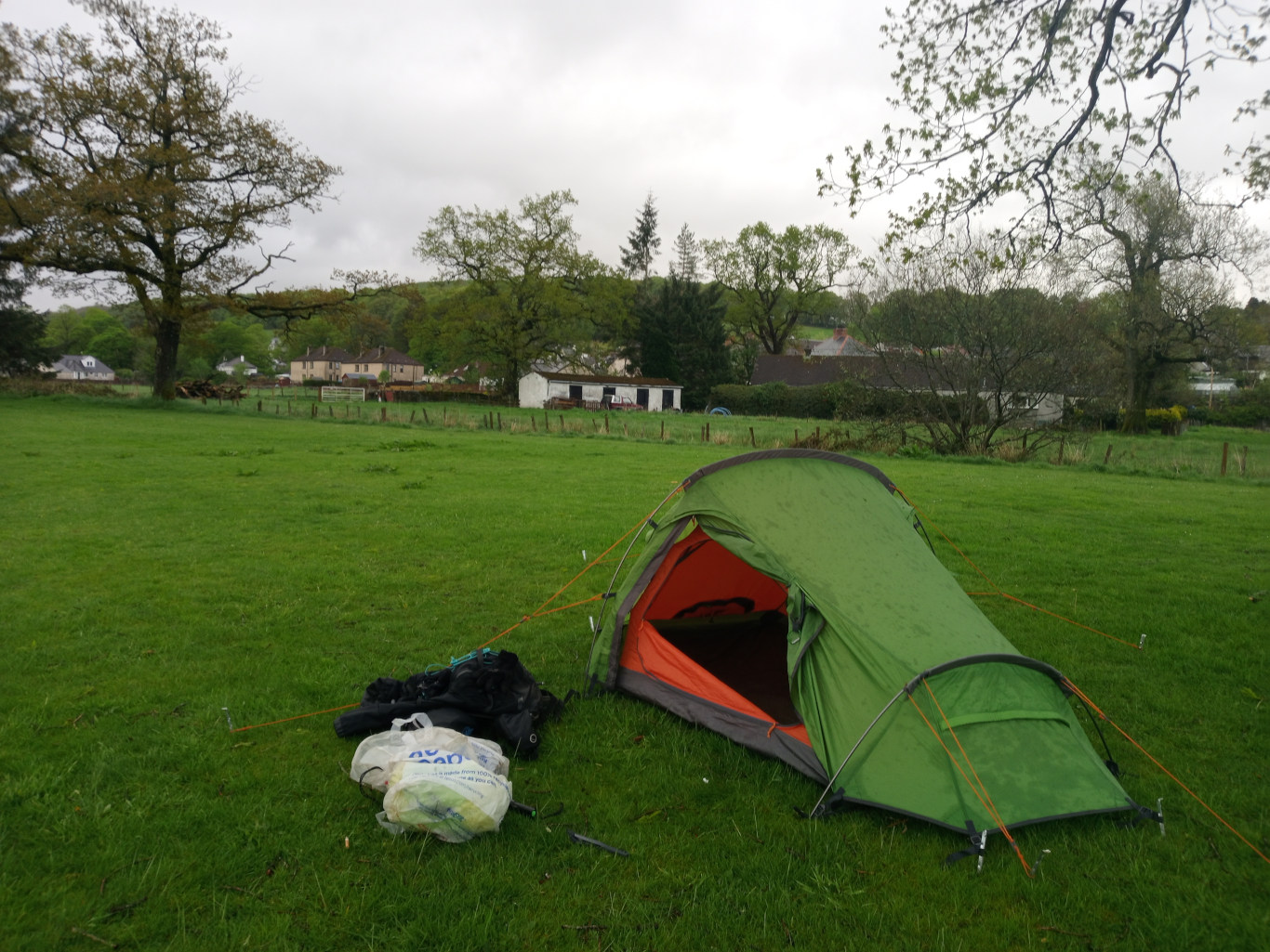 Central focus is a bright green, dome-shaped one-person tent pitched on a lush green field. The tent's inner lining is visible through the open doorway, revealing a vibrant orange interior. It's slightly damp, showing subtle dark spots from recent rain. Orange guy lines extend from the tent, securing it to the ground with pegs. Beside the tent, a pile of camping gear rests on the grass; a black backpack, possibly a sleeping bag, and a crumpled, light blue and white plastic grocery bag are discernible. A few tent pegs and a hiking pole lie scattered near the gear. The scene is set in a pastoral landscape under a cloudy, overcast sky. The light is diffuse and soft, indicating an absence of harsh shadows. The predominant color is the vibrant green of the grassy field, which extends to the horizon. In the background, a row of modest houses with grey and brown roofs is visible, nestled amongst a cluster of mature deciduous trees exhibiting varying shades of green and a few touches of lighter yellow-green indicating new leaves.