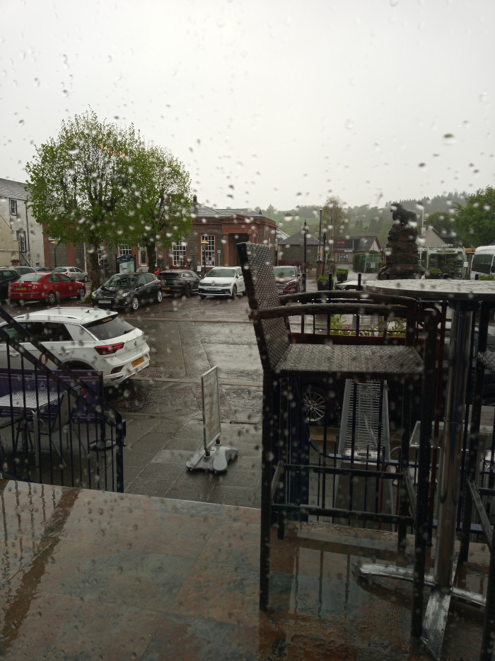 Inside a building, looking out through a rain-streaked window. The focus is on a wet, paved area outside with parked cars. The foreground is dominated by the out-of-focus image of patio furniture – a dark-colored wicker chair and metal table set. These appear wet from the rain, and the rain droplets distort their appearance. Part of a metal frame or stand also occupies the foreground. A small, grey A-frame sign stand is near the base of the window. A staircase is partly visible to the left of the furniture.