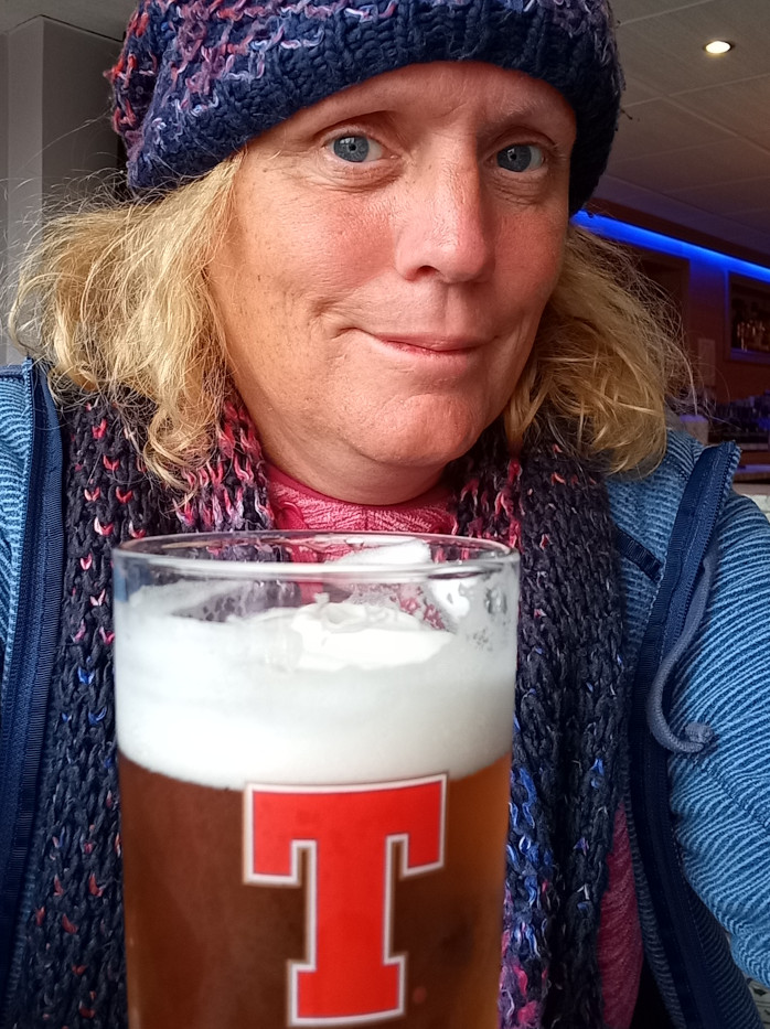 Enters on a middle-aged woman with shoulder-length, light blonde hair. She's wearing a dark blue and purple knitted beanie pulled low on her forehead, obscuring some of her hairline. A chunky, multicolored knitted scarf, predominantly dark blue, purple, and hints of pink, is wrapped around her neck. She is dressed in a blue zip-up jacket. Her expression is neutral, almost pensive, with a slight downward curve of her lips, creating a mildly melancholic yet calm mood. The woman is holding a pint glass of beer that appears to be a light-colored ale with a thick, creamy white head. The glass prominently displays a large red T logo, indicating a specific brand. She's slightly turned toward the viewer, creating a close-up intimate perspective.