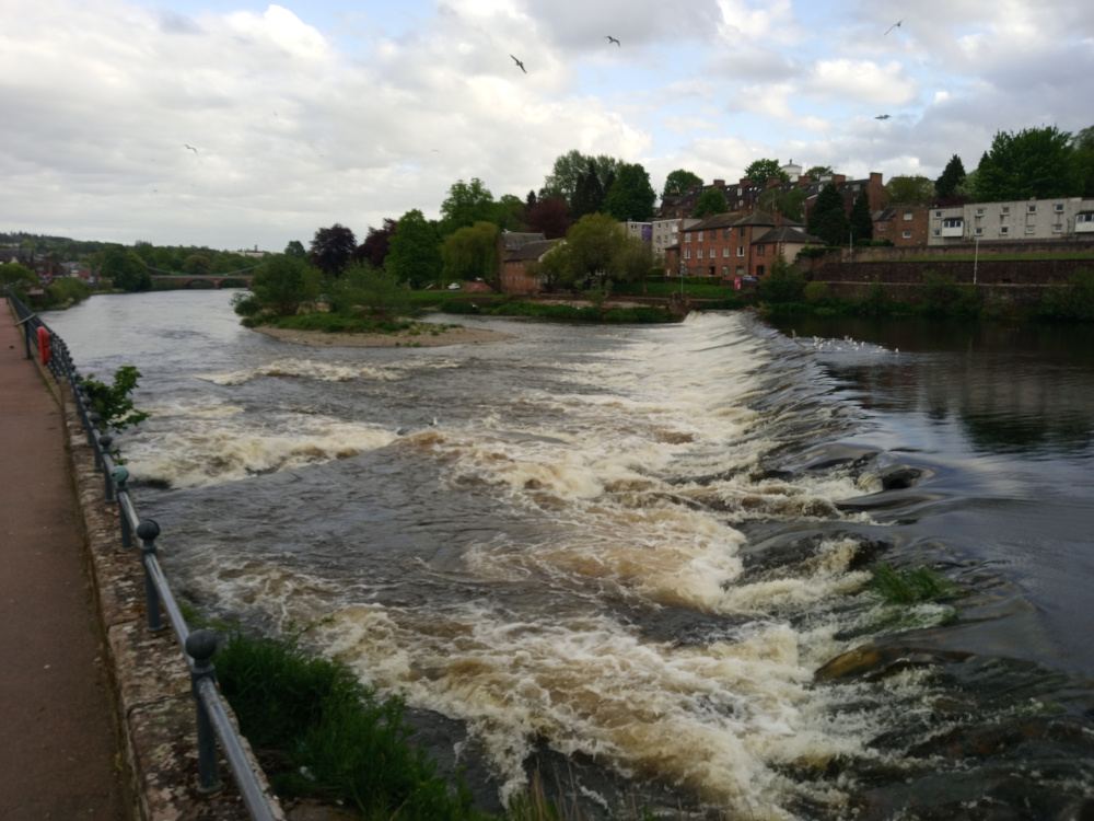 Rushing river, its water a churning mix of dark brown and frothy beige, cascading over a low, stone weir that spans most of the image's width. The water's movement is dynamic, with frothy whitecaps and swirling currents visible throughout. A metal railing, dark grey with rounded finials, runs along the left edge of the image, paralleling a red-brown paved pathway. Sparse, low-lying vegetation clings to the riverbank near the railing. In the background, a line of brick buildings, a mix of residential houses and what appears to be an apartment complex, rises on a slightly elevated bank. Trees, primarily green and lush with hints of yellow-green, line the riverbank and fill the spaces between the buildings. A few birds are visible in the sky, small against the vastness of the clouds. There's a suggestion of a bridge in the far background spanning the calmer river section before the weir.