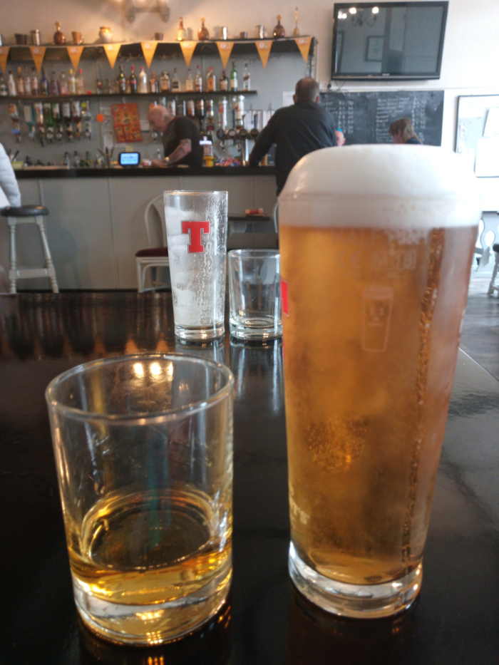 Dark, polished table in a pub setting, showcasing a collection of glassware. Dominating the foreground are: a short, squat tumbler partially filled with a golden amber liquid (likely whiskey or a similar spirit), positioned slightly off-center to the left; and a tall pint glass brimming with a light, golden-hued beer, topped with a thick, frothy head, situated to the right. Between these two, slightly blurred and out of focus, is a smaller, empty glass. Behind them, a taller, frosted pint glass, bearing a red T logo (suggesting a specific brand of beer), sits on the table's surface. In the background, blurred but discernible, are patrons seated at a bar. A man with short, dark hair is visible in the center, wearing a dark jacket, seemingly engrossed in conversation. To his left, another man with a shaved head and a dark shirt is seen from behind, partially obscured, seemingly involved in a task behind the bar. A woman with lighter hair is partially visible to the right, possibly also sitting at the bar. A muted, darker figure is partly shown in the very far left corner. The setting is a traditional pub with a warmly lit interior. The background shows a long bar stocked with a vast array of liquor bottles neatly organized on shelves. These bottles shimmer under the lighting, creating a sense of depth. Behind the bar, there is a television screen displaying a dark image, adding a layer of subtle contrast. A dark, possibly chalkboard, menu board is partially visible on the back wall. The walls are a light, neutral color, offset by the dark wood of the bar and the darker, polished tabletop. The lighting is softly diffused, casting a warm, inviting ambiance, with subtle highlights reflecting off the surfaces of the glasses.