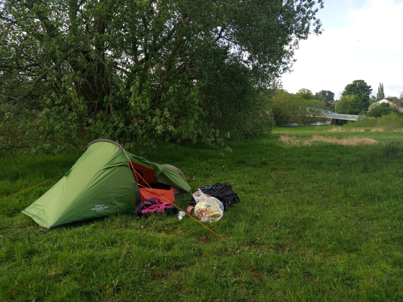 Vibrant green, one-person backpacking tent, a Banshee 200, pitched on a lush, grassy field. It's slightly angled to the left, its entrance partially open, revealing a glimpse of an orange inner tent. Beside the tent, on the grass, lies a haphazard collection of camping gear: a partially visible backpack, a clear plastic bag containing what appears to be clothing or supplies, and other smaller items, possibly toiletries or cooking utensils, scattered around. The scene suggests a temporary campsite. There are no people present.