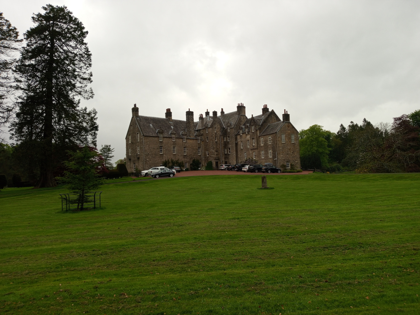 Central focus is a large, imposing stone mansion, typical of a Scottish country house or castle. It's a multi-story structure with multiple gables, numerous windows, and a grey-brown stone exterior. Several cars are parked in a line in front of the building, suggesting a driveway or parking area. A small, dark grey car is prominently positioned slightly off-center, just in front of the mansion. Other cars are less distinct. In the foreground, a lush, vibrant green lawn stretches across the entire lower half of the image. A simple, black metal garden structure, possibly a small trellis or plant stand, stands alone on the left side of the lawn, mid-ground. A lone, relatively small deciduous tree stands near the garden structure, creating contrast in scale and texture with the tall conifer in the upper left.
