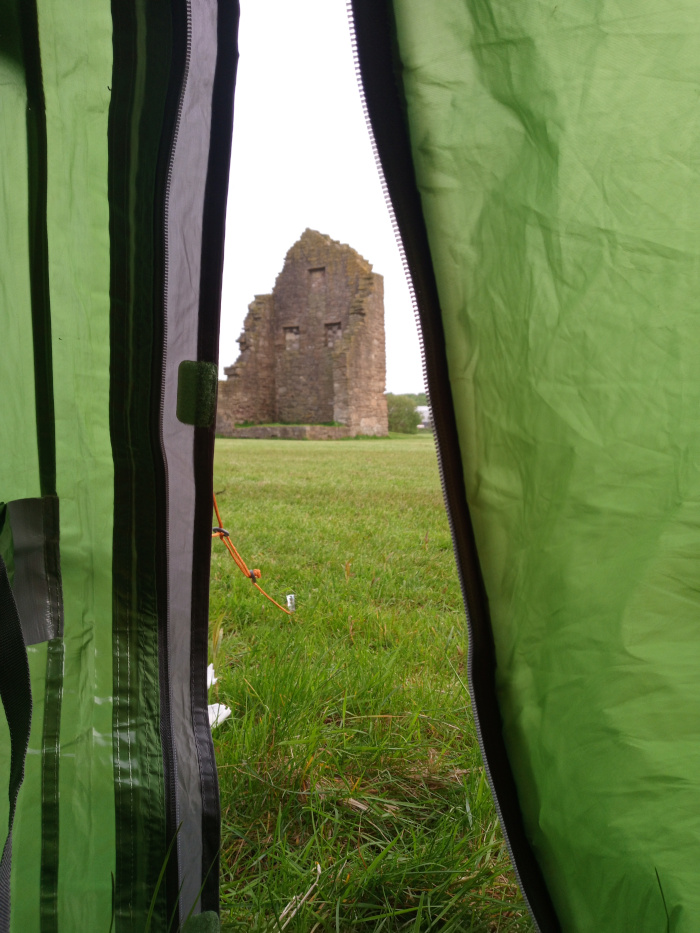 Predominantly a view from inside a green tent, specifically from between two slightly ajar sections of the tent's fabric. The tent's material is a vibrant, slightly crumpled, lime-green nylon-like fabric with darker grey zipper seams running vertically. A small, light grey plastic piece is visible near the upper-left side of the grey seam. An orange-brown cord is partially visible, extending from inside the tent towards the outside. The main focus is on the view through the tent opening, which reveals a stone ruin in the mid-ground. The ruin is a substantial, grey stone structure with visible holes that suggest windows or former openings. It's in a state of disrepair, with portions visibly missing and showing its age. The background beyond the ruin consists of a field of tall, lush, vibrant green grass. No people are visible in the image.