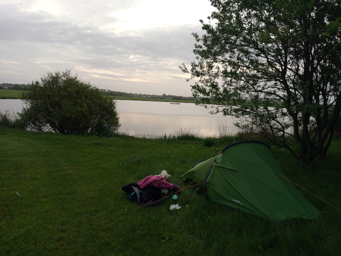 Vibrant green, single-person tent pitched on a grassy bank. It's slightly angled, its entrance facing away from the viewer toward a calm body of water. Near the tent, a pile of camping gear is visible, including a dark-colored backpack and a purplish-pink item that appears to be clothing. Some crumpled white trash or tissue is scattered near the gear. There is no human figure directly visible, but a pile of clothing near the tent suggests a person has recently been there. The scene unfolds at the edge of a tranquil lake or pond. The water is relatively still, reflecting a muted sky. The far bank of the water is low and mostly flat, with a line of trees and shrubs creating a soft, horizon line. The sky above is overcast, with soft, diffused light that lacks harsh shadows.