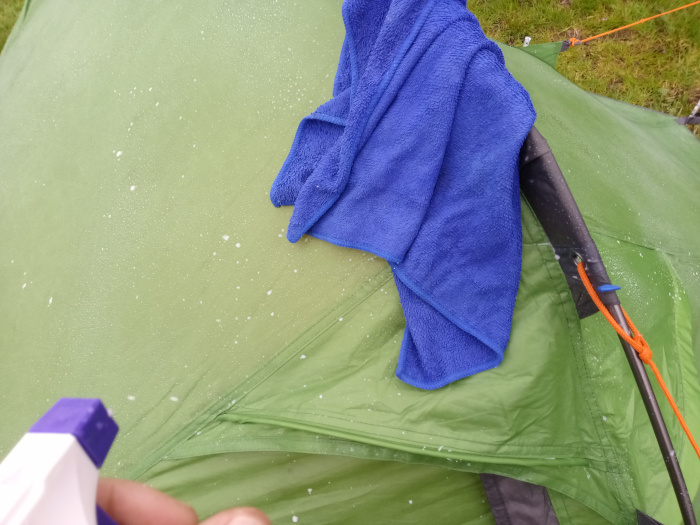 Leonie's hand, partially visible in the bottom left corner, holding a white spray bottle with a purple nozzle. This bottle is pointed towards a green tent's surface. The tent fabric is visibly damp, speckled with small, white droplets—likely from a cleaning solution recently sprayed. A vibrant blue microfiber towel, loosely folded and slightly rumpled, rests atop the tent's fabric near the top centre. The towel shows a slightly damp appearance, suggesting it's been used in the cleaning process. The tent itself is olive-green, with darker gray poles and bright orange cords visible. The tent fabric shows minor creases and wrinkles. The tent's structure includes seams and a distinct pole structure visible.