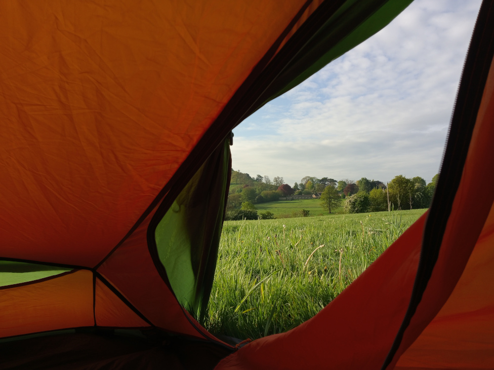 Interior of a brightly coloured, orange and green dome tent. The tent fabric is slightly wrinkled and shows the texture of nylon or a similar material. A section of the tent's zip is visible, suggesting the tent is partially opened. No people or other objects are inside the tent. The background seen through the opened section of the tent is a picturesque pastoral scene. A field of vibrant, spring-green grass stretches from the bottom edge of the frame to a line of low-lying hills in the distance. On these hills, we see a cluster of quaint buildings — possibly a small village or farmstead — with a scattering of trees, creating a naturalistic, bucolic landscape. The sky is a soft, pale blue, lightly clouded, with hints of white and grey, suggesting a calm, possibly early morning or late afternoon light.