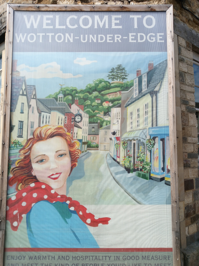 Cheerful, rosy-cheeked woman with shoulder-length auburn hair styled in soft waves. She's positioned slightly off-centre, facing the viewer but angled slightly to her left. She wears a light teal, almost turquoise, dress and a vibrant red polka-dotted scarf loosely draped around her neck and shoulders. Her expression is welcoming and friendly, with a gentle smile. She's not actively doing anything; her pose suggests a relaxed, inviting presence. The background features a charming depiction of a quaint village street, Wotton-under-Edge, nestled amongst rolling green hills. Buildings are rendered in various shades of warm yellows, creams, and muted browns, suggesting aged stone and timber construction. Small details, like window boxes overflowing with flowers and shop signs, add to the village’s lived-in quality.