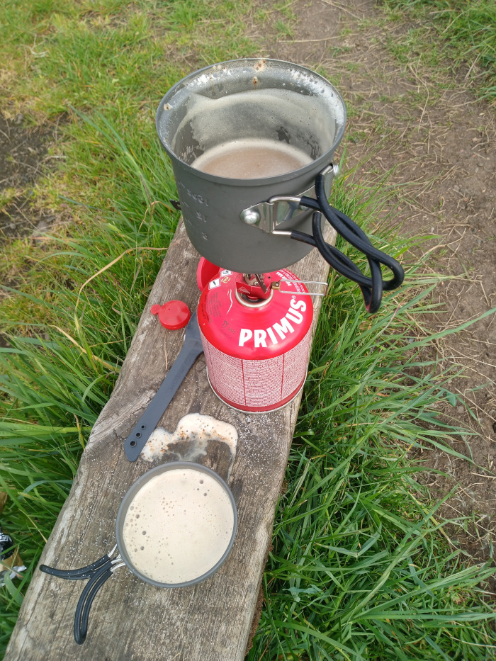Rustic outdoor scene depicting the preparation of a hot beverage, likely coffee or tea, using a portable camping stove. Dominating the foreground is a weathered, gray-brown wooden bench or log. Resting on it are two dark gray metal camping mugs. The top mug, larger, sits atop a Primus brand red canister stove; it's filled with a light brown, frothy liquid, suggesting recently boiled or brewed beverage. The second mug, smaller, sits below and to the right, also containing a similar light brown, frothy liquid. A dark gray, spatula-like utensil lies next to the stove and a small red cap is near the stove's base.