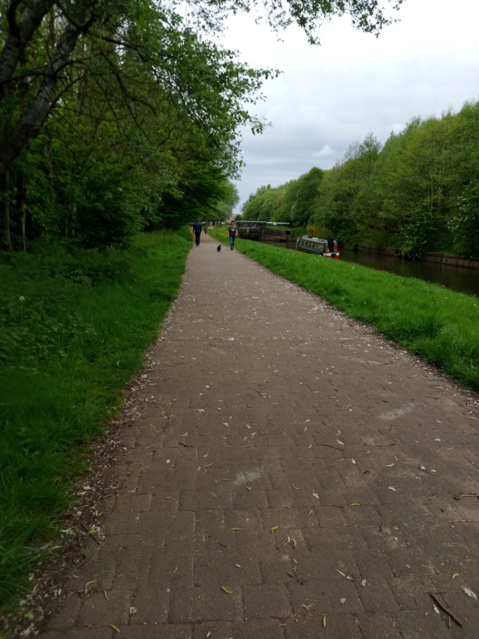 Paved towpath, light brown-grey in colour, composed of slightly irregular, rectangular paving stones. Scattered across the path are numerous small, light-coloured debris, possibly dried flower petals or small twigs. Two figures are visible in the mid-ground, walking away from the viewer towards a canal lock. One appears to be wearing darker clothing, the other lighter. A small, dark-coloured dog is walking between them. Further in the background, a narrowboat (a long, narrow boat used on canals) is moored on the canal.