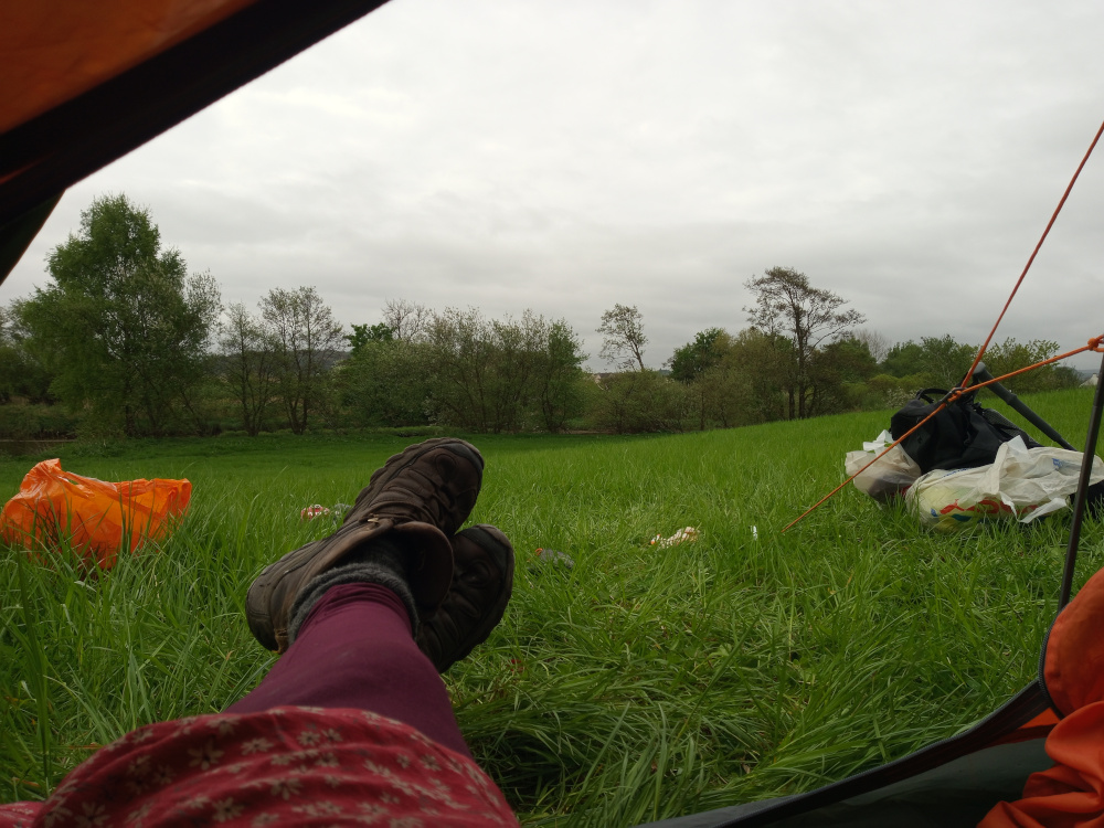 First-person perspective shot from inside a tent. The viewer's legs, clad in maroon tights and a red, floral-patterned dress or skirt, are prominently displayed, extending from the bottom of the frame towards the top. The person is comfortably reclined with their feet propped up. The feet are shod in dark brown hiking boots. One boot sits angled slightly higher than the other. Outside the tent, in the foreground, lies crumpled orange and plastic bags. A dark backpack and other pieces of luggage rest further away, slightly to the right. The scene is set in a lush green field, under a muted, overcast sky. The grass is vibrant green, slightly long, appearing slightly damp. The lighting is soft and diffused, characteristic of a cloudy day. The sky is a consistent light gray-white, devoid of intense brightness or shadows. Behind the field, a line of deciduous trees runs horizontally across the image, forming a natural border between the field and a more distant, indistinct landscape. The trees are a muted green and brown, lacking any vibrant summer hues.