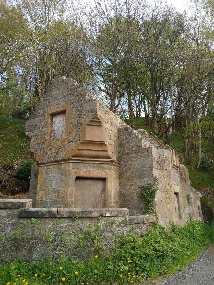 Crumbling, stone structure, possibly a ruin of a small building or outbuilding. Its walls are made of weathered, light-brown and tan stones, unevenly stacked, showing significant signs of age and decay. Some sections of the walls are missing entirely, revealing the interior. One visible wall features a recessed window, now lacking glass, and a boarded-up doorway. The structure has a slightly angled, almost asymmetrical, shape, emphasising its decay. A low stone wall runs horizontally across the foreground, partially obscuring the base of the ruin. This wall is also made of similar stone but appears in better condition than the main building. The scene is set in a natural environment, possibly a hillside. Behind the ruin stands a dense stand of deciduous trees, their leaves light green and partially obscuring a hillside, indicating springtime. The trees’ branches create a backdrop of intricate lines and shadows. The hillside behind the trees appears grassy and gently sloping. The overall lighting is soft and diffuse, suggesting an overcast day or soft sunlight filtering through the trees.
