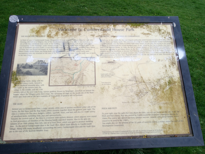 Weathered, rectangular information board, dark brown-framed, situated outdoors on a short, grassy post. The board is predominantly off-white or beige, heavily speckled with dark brown stains and patches of what appears to be lichen or mold, especially at the edges. The text is printed in dark brown, a serif font, and includes historical information about Cumbernauld House Park, along with small historical maps and illustrations. The illustrations are simple line drawings, somewhat faded and showing age. There's some evidence of graffiti or vandalism – light splotches of white, possibly paint, mar the surface. No people are present.