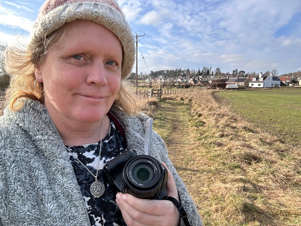 leonie with her bridge camera by the ale water, ancrum is seen in the distance behind her