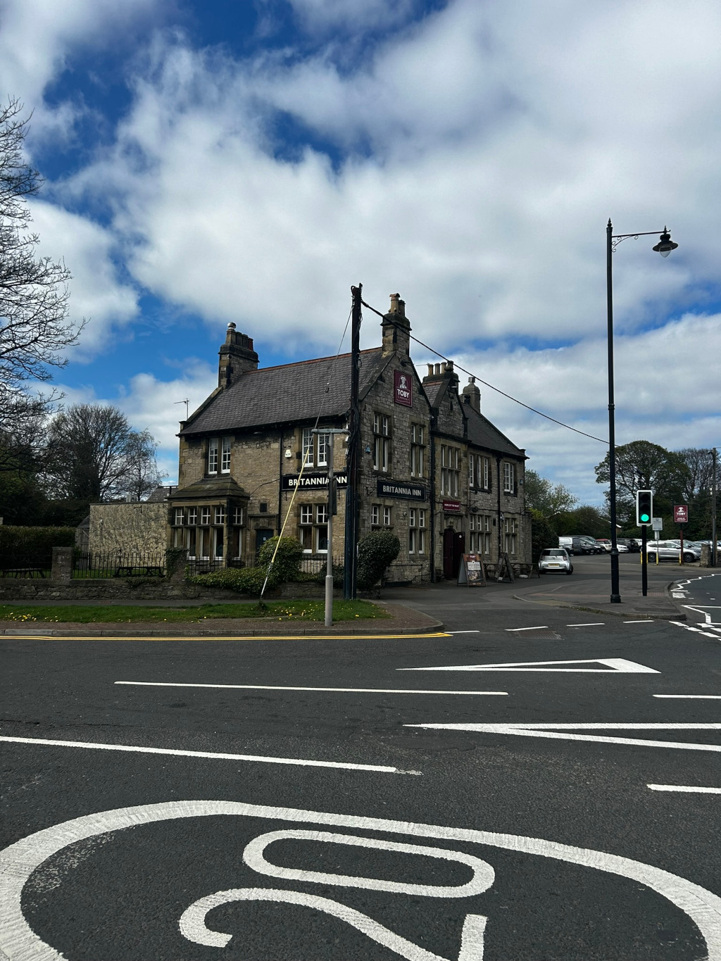 Britannia Inn, a stone pub, situated on a corner where two roads intersect. The pub is a two-story building with traditional architecture. The sky is partly cloudy, with a mix of blue sky and fluffy white clouds. There are cars parked nearby, and a traffic light is visible at the intersection. The overall scene suggests a quiet, possibly suburban, location in the UK.