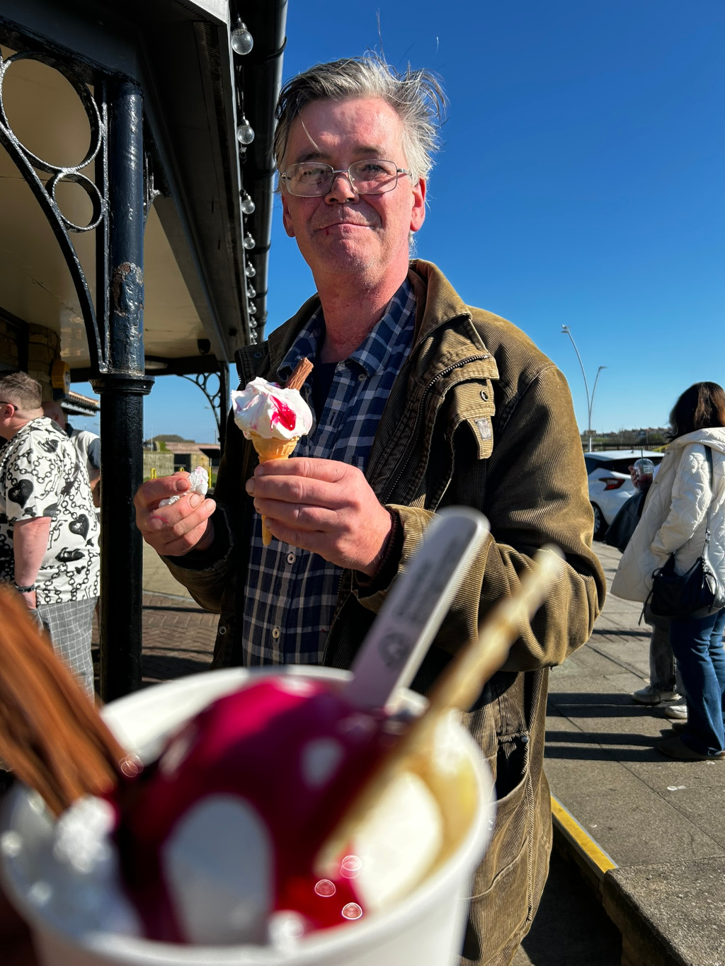 Charlie with glasses and slightly windblown gray hair, smiling at the camera while holding an ice cream cone in his hands. He is wearing a brown corduroy jacket over a plaid shirt. In the foreground, out of focus, is another ice cream dessert in a cup. The background features a seaside promenade setting, with other people visible in the distance, suggesting a sunny and pleasant day. The overall impression is one of casual enjoyment and relaxed happiness.