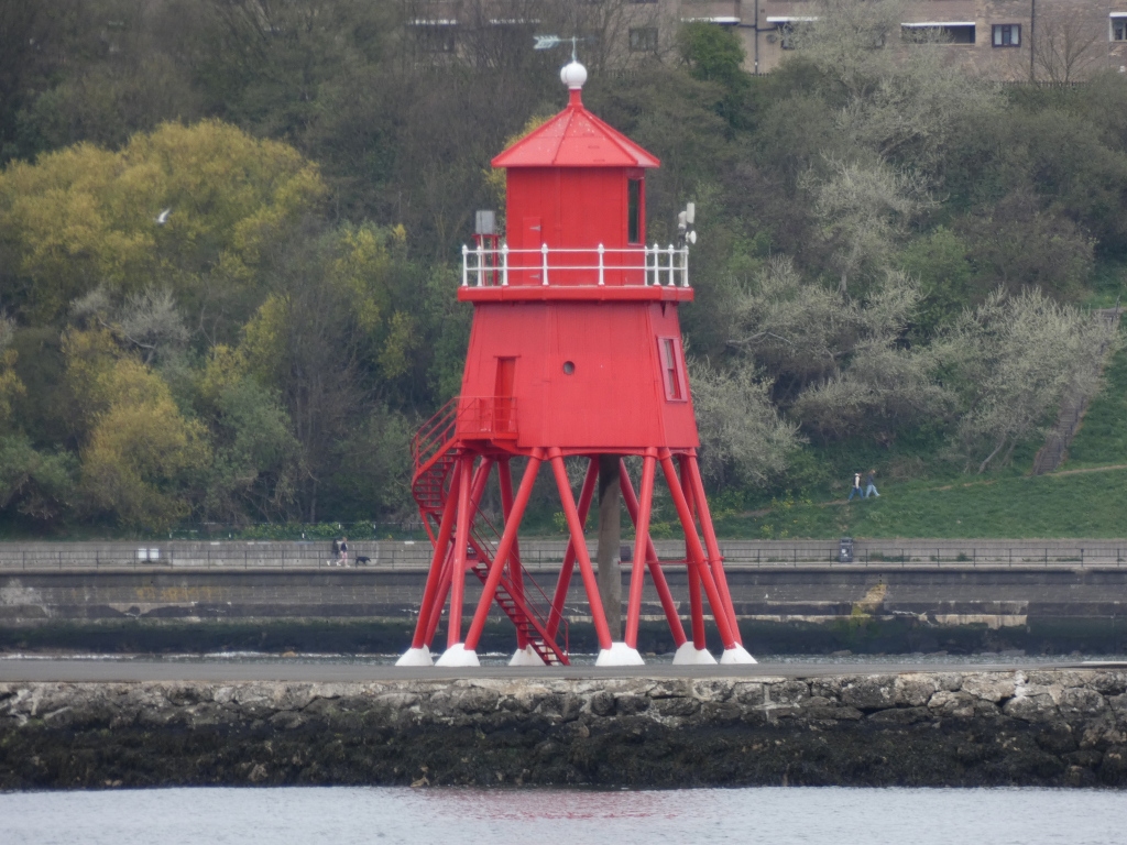 Bright red lighthouse standing on stilts in a body of water. It's a relatively small structure with a white railing and a spiral staircase on the outside. Behind the lighthouse is a low, rocky seawall and a background of trees and a distant building. A few people are visible walking along the seawall in the distance.
