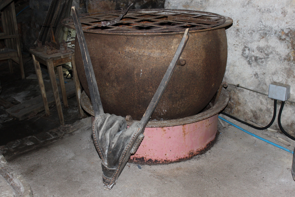Large, rusty cauldron sitting on a smaller, pink metal base. A bellows, appearing old and worn, is positioned in front of the cauldron, suggesting its use in fuelling a fire beneath. The setting appears to be a dark, possibly old, basement or cellar, contributing to an overall atmosphere of age and disuse. The scene evokes a feeling of history and forgotten crafts.