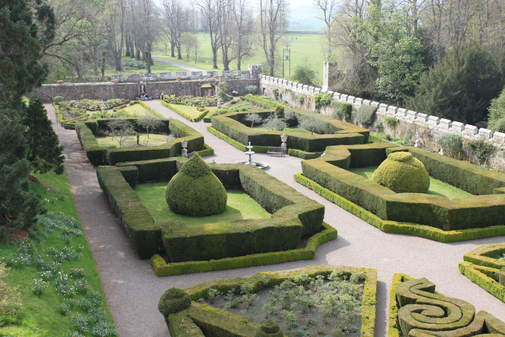 Meticulously designed formal garden, viewed from an elevated perspective. The garden features intricately shaped hedges forming geometric patterns and compartments. There are several topiary plants, including large, rounded ones, adding to the structured aesthetic. A gravel pathway winds through the garden, connecting different sections. In the background, a stone wall and the tops of trees are visible, suggesting the garden is part of a larger estate or castle. A few people can be seen in the distance near the wall, providing a sense of scale.