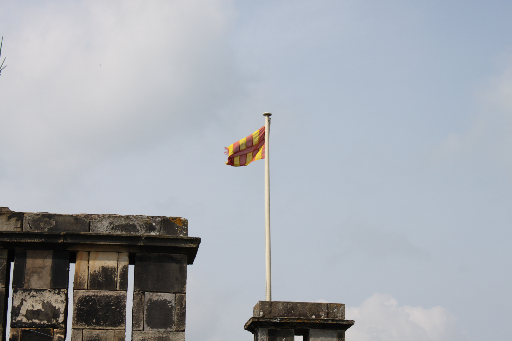 Flag with red and yellow horizontal stripes waving on a flagpole against a cloudy sky. The flagpole is mounted on a small stone structure, and in the foreground to the left is a portion of a larger, weathered stone structure, possibly part of a wall or ruin. The overall mood is quiet and somewhat melancholic.