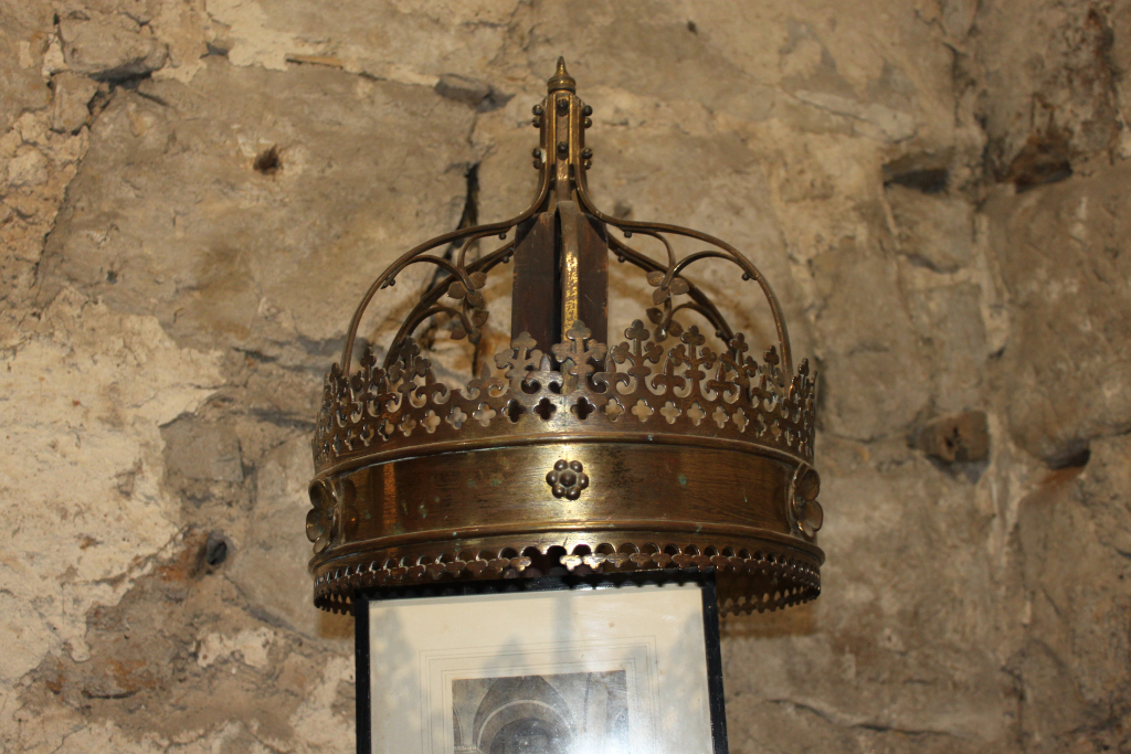 Close-up view of an ornate, crown-shaped brass lamp or light fixture. It's affixed to what appears to be a dark-Coloured wooden or metal post, which in turn is set against a rough, textured stone wall. The crown features intricate detailing, including floral and leaf-like motifs, and is topped with a pointed spire. A partially visible picture or print is underneath the crown within the lamp's housing.