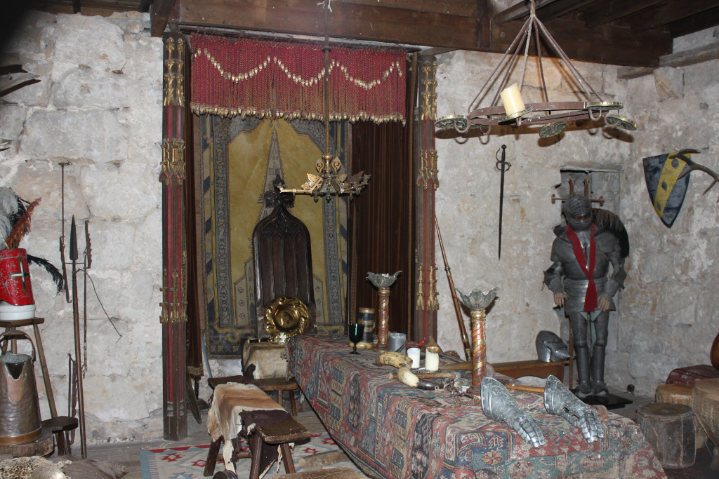 Interior of a medieval-style room, possibly a castle chamber. The focal point is a long, richly decorated table laden with various medieval-style objects, including candlesticks, goblets, and what appear to be gauntlets. Behind the table is an ornate chair, a tapestry, and a suit of armour stands against the stone wall, along with various weapons and other medieval artefacts.