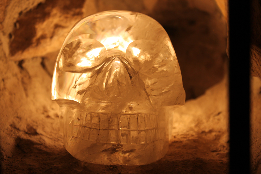 Crystalline skull, seemingly carved from quartz or a similar translucent material. It’s positioned within a dimly lit, earthy alcove, giving the impression of a relic or artifact found in a cave or ancient structure. The light source, not directly visible, illuminates the skull from within, enhancing its translucence and creating a warm, almost ethereal glow. The overall effect is one of mystery and perhaps even a touch of the macabre, contrasting the natural, rough texture of the surrounding stone with the smooth, precisely carved skull.