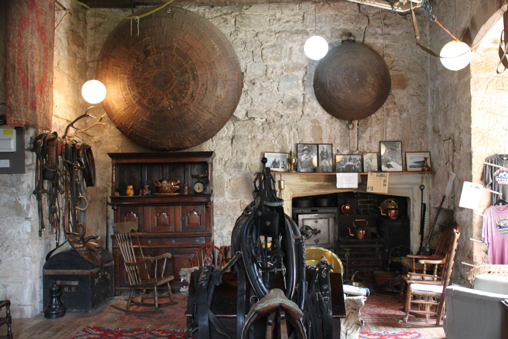 Interior of a rustic, stone-walled room, possibly in a historic building. It's filled with antique furniture and various items, including two large, old metal serving platters hanging on the wall, a wooden rocking chair, an old wooden chest, and a collection of antique harnesses and saddles. The room also includes an old fireplace with copper pots, and several framed photographs on the mantelpiece.