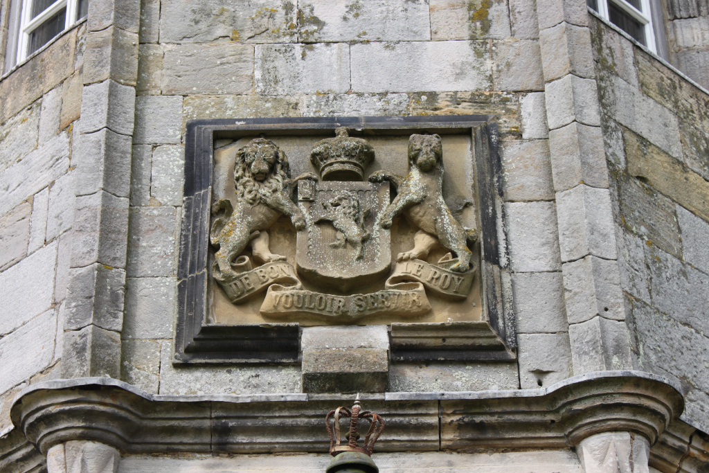 Stone carving of a coat of arms, set into the stonework of a building. The coat of arms features two lions rampant supporting a shield, with a crown above. A scroll at the bottom bears an inscription. The stonework shows signs of age and weathering. Below the coat of arms is a smaller, separate stone crown. The overall impression is of an aged, historical building detail.