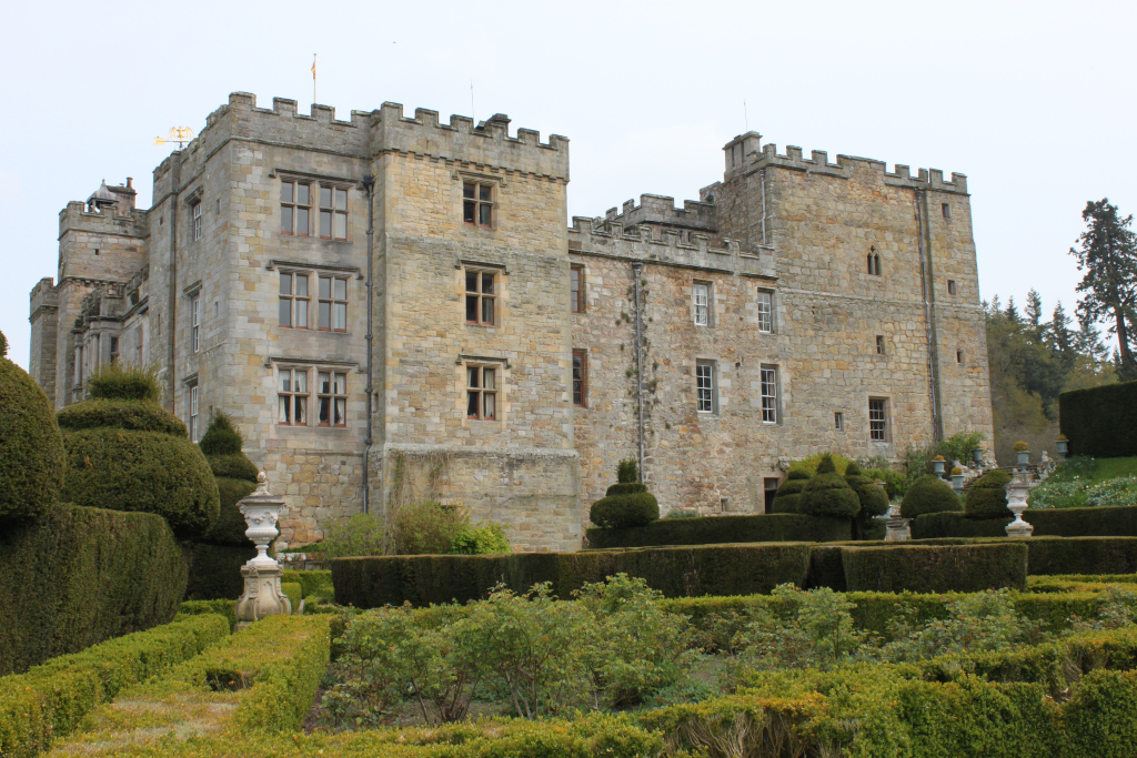 Large, imposing stone castle, possibly of medieval or Tudor origin, set within meticulously maintained formal gardens. The castle’s exterior is made of light brown stone, showing its age and exhibiting a crenelated battlement on top of its walls. The gardens feature neatly trimmed hedges forming geometric patterns, interspersed with ornamental stone urns. The overall impression is one of wealth, history, and carefully cultivated beauty.