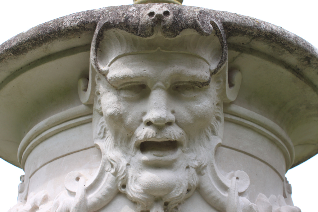 Close-up of a weathered stone carving of a bearded man's face. The face is centrally positioned and framed by ornate stonework that appears to be part of a larger structure, possibly a fountain or column. The man's expression is serious, almost stern, with his mouth slightly open. The stone shows signs of age and weathering, adding to its historical and possibly classical feel.