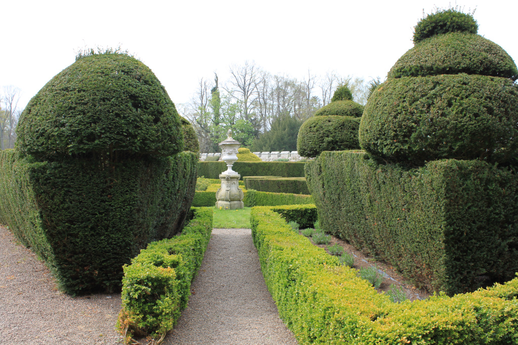 Formal garden with a gravel path winding through meticulously manicured hedges. The hedges are shaped into geometric forms, including large, rounded balls and rectangular sections. A stone urn sits on a pedestal in the middle ground, adding a focal point to the scene. The background includes some deciduous trees, suggesting a park-like setting. The overall impression is one of order, symmetry, and careful landscaping, characteristic of a formal European garden style.