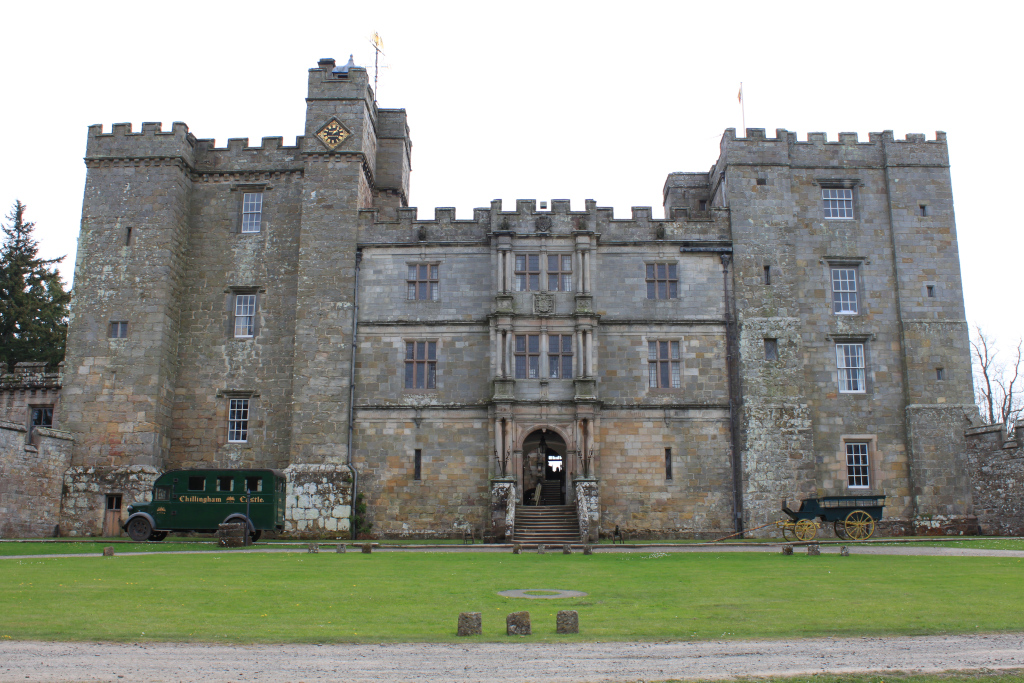 Full frontal view of Chillingham Castle in Northumberland, England. The castle is a large, imposing stone structure with crenellations, multiple towers, and numerous windows. In the foreground is a neatly kept lawn, and parked near the castle are a vintage green bus and a horse-drawn carriage, suggesting a perhaps tourist-oriented setting.