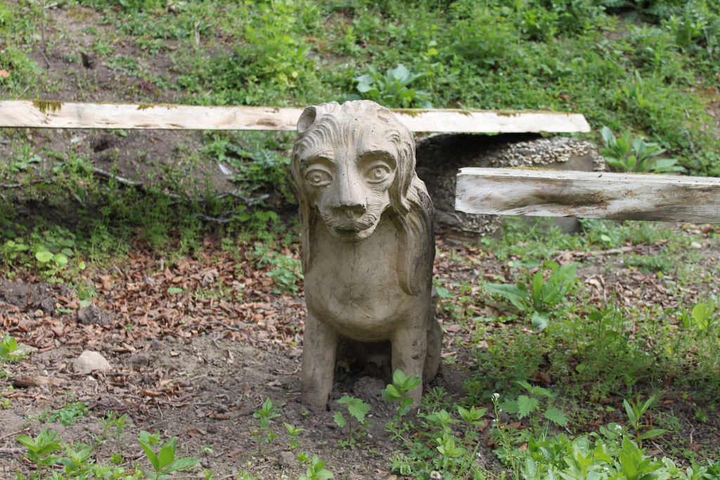 Weathered stone statue of a dog sitting on the ground amidst overgrown vegetation. The statue appears to be old and possibly part of a garden or park setting. Parts of weathered wooden planks are visible in the background.