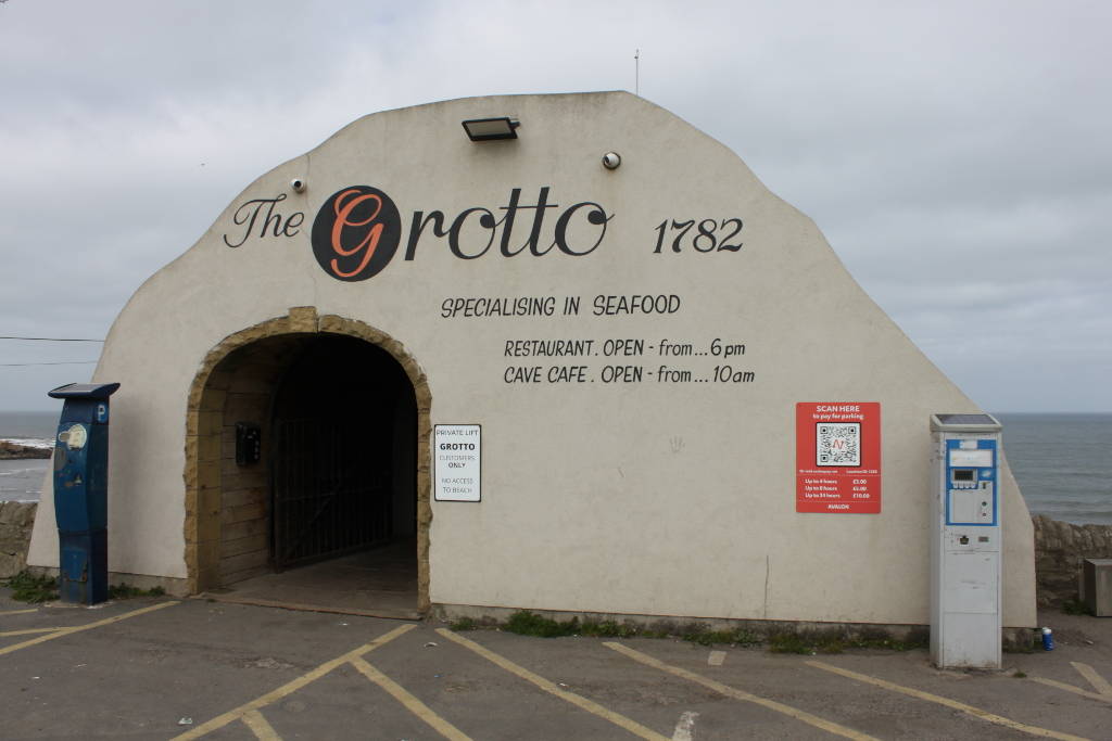 The entrance to a seafood restaurant and cafe called The Grotto, established in 1782. The building is styled to resemble a cave entrance. Signage indicates the restaurant and cafe's operating hours, and smaller signs indicate a private lift for customers only and a parking payment system using a QR code. The location is coastal, with a view of the ocean visible in the background. The overall impression is one of a quaint, seaside establishment.