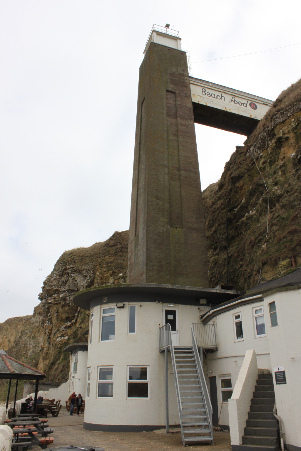 Tall, narrow, grey stone tower attached to a white, cylindrical building. A walkway, labelled Beach Food, bridges the gap between the tower and a cliff face. There are stairs leading up to the building's entrance. In the foreground, there are outdoor tables and chairs, suggesting a cafe or restaurant setting. The overall setting appears coastal.