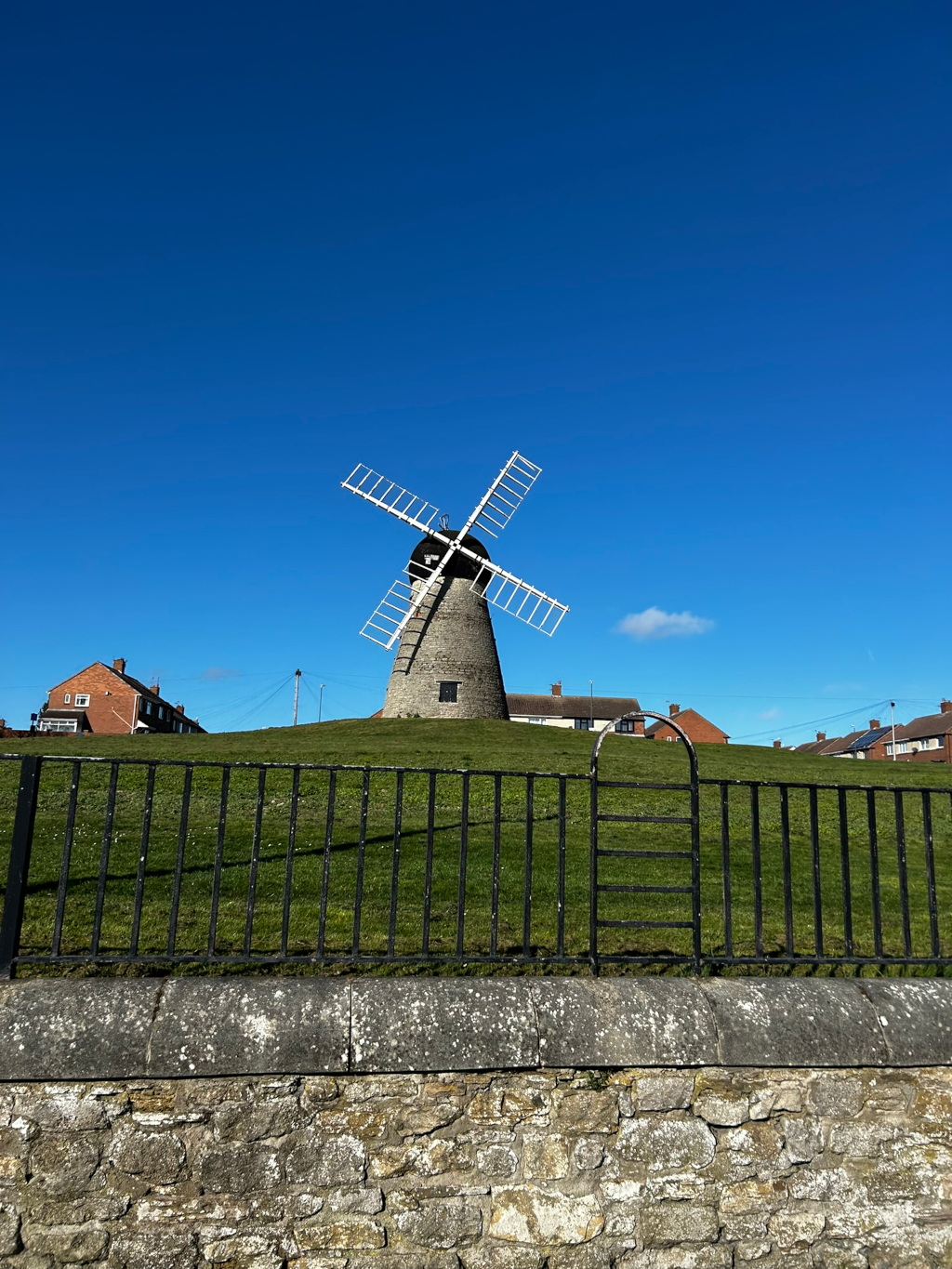 A stone windmill with white sails set against a clear blue sky. The windmill sits atop a grassy hill, behind a black metal fence and a low stone wall. Residential houses can be seen in the background. The overall impression is one of a peaceful, idyllic setting.