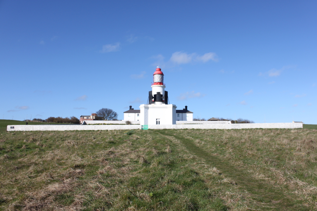 Lighthouse situated atop a grassy hill. The lighthouse is predominantly white with a red lantern room at the top. It's set against a clear, bright blue sky, with a low wall surrounding its base and some buildings connected to it. A grassy path leads towards the lighthouse from the foreground. The overall scene is peaceful and evokes a sense of coastal serenity.