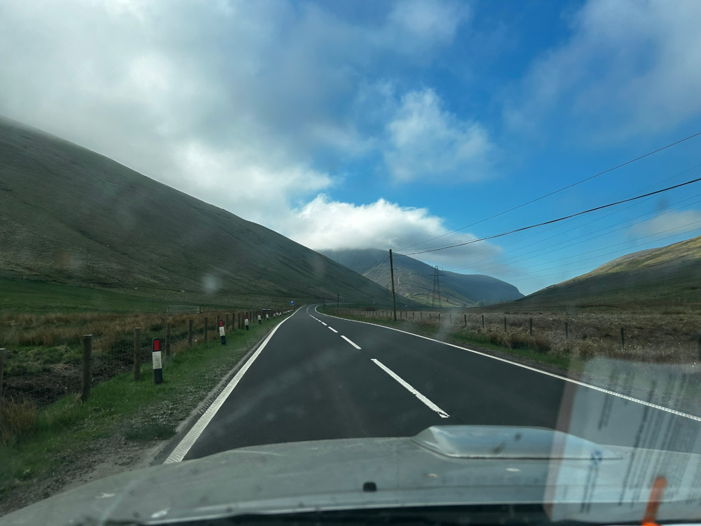 View from inside a vehicle driving along a winding road in a mountainous region. The road is flanked by verdant hillsides, and the sky is mostly clear blue with some fluffy white clouds, one of which partially obscures a mountain peak in the distance. There are power lines visible running along the roadside. The overall impression is one of a scenic, possibly remote, drive through open countryside.
