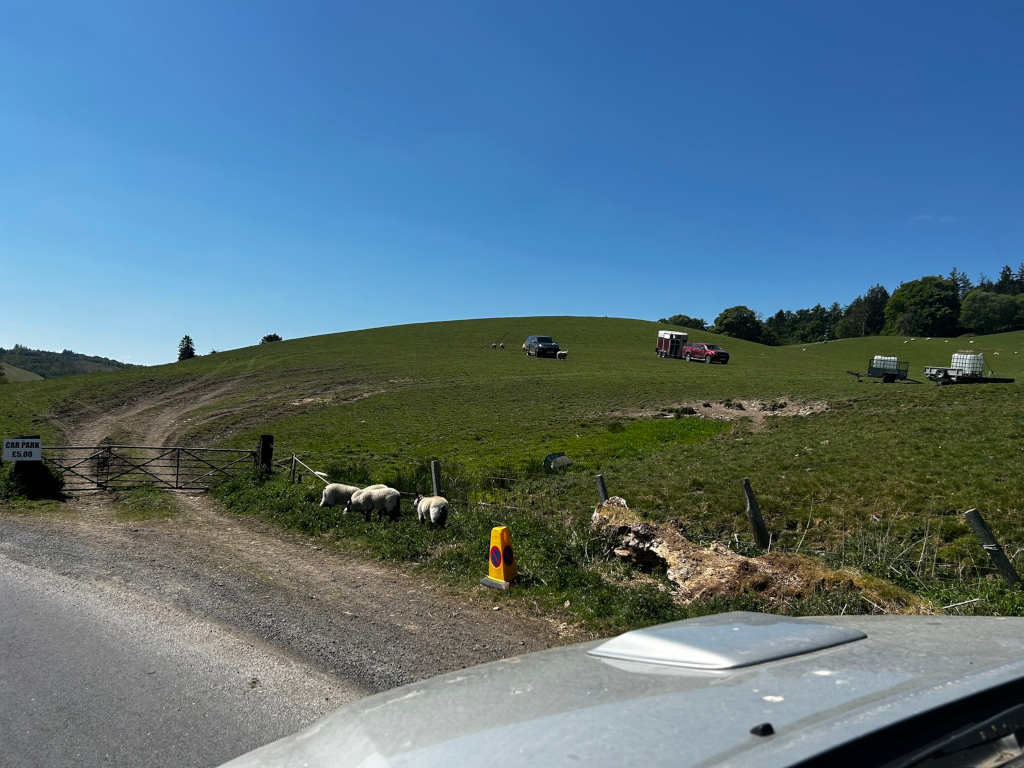 Grassy hillside on a sunny day. In the middle ground, there are a couple of vehicles, one appearing to be a horse trailer, and a few sheep grazing on the hill. There's a car park sign visible near the bottom left, indicating what appears to be a rural or countryside location. A road or track leads towards the hill's base. The foreground shows a part of a car's roof and windshield, suggesting that the photo was taken from inside a vehicle. The overall scene is peaceful and pastoral.
