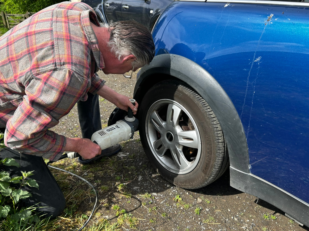 Charlie kneeling beside a blue Mini Cooper car. He is using a power tool, possibly a grinder or polisher, on the wheel arch. Charlie is wearing a plaid shirt and appears to be performing some kind of car maintenance or repair. The setting appears to be outdoors.